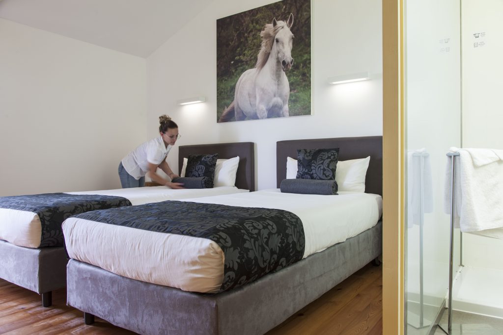 Hotelkamer met twee bedden en vrouw die de kussens recht legt. Boven het bed hangt een foto van een paard.